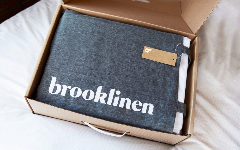 Brooklinen plain kraft corrugated shipper opened to reveal linens in a branded canvas dust bag with hang tag